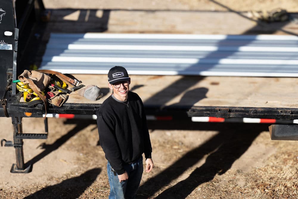 Smiling man in a black shirt and cap stands by a trailer with construction materials.